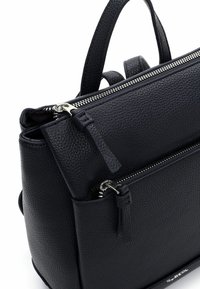 Czarna torebka ze skóry z fakturą, z zamkiem błyskawicznym u góry, przednią kieszenią, metalowymi elementami i dwoma krótkimi uchwytami. Kompaktowa o wyraźnej, usztywnionej formie.