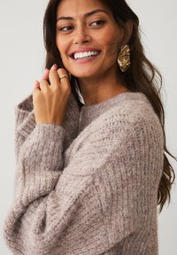 Lys beige strikket sweater med et tekstureret mønster, brede ærmer og en løs pasform. Gyldne statement øreringe komplimenterer looket.