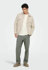 Veste beige avec deux poches poitrine, portée sur une chemise blanche. Pantalon gris à texture lisse ; chaussures marron clair avec une semelle épaisse.