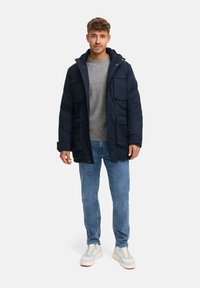 Marineblå dunjakke med flere frontlommer, grå striktrøje, lyseblå jeans og hvide sneakers med grå detaljer.