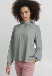 Ljusgrön långärmad blus med hög halsringning, invecklad detalj fram och elastiska manschetter, kombinerad med rosa byxor.