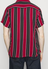 Mężczyzna w koszuli z krótkim rękawem w pionowe czerwono-czarno-białe paski, w ciemnoniebieskich dżinsach, pokazany od tyłu.