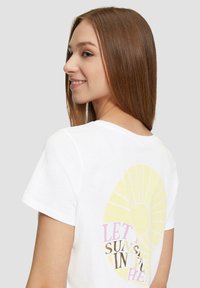 Witte katoenen t-shirt met een grote gele zonafbeelding en roze en gouden tekst "Laat de zonneschijn in je." Korte mouwen en een relaxte fit.