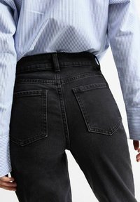 Persona che indossa jeans neri in denim a vita alta e una camicia a maniche lunghe blu e bianca a righe, vista da dietro.