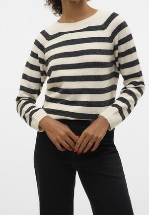 Kobieta w kremowo-czarnym swetrze w poziome pasy z długimi rękawami i czarnych spodniach, z rękami złożonymi na wysokości talii.