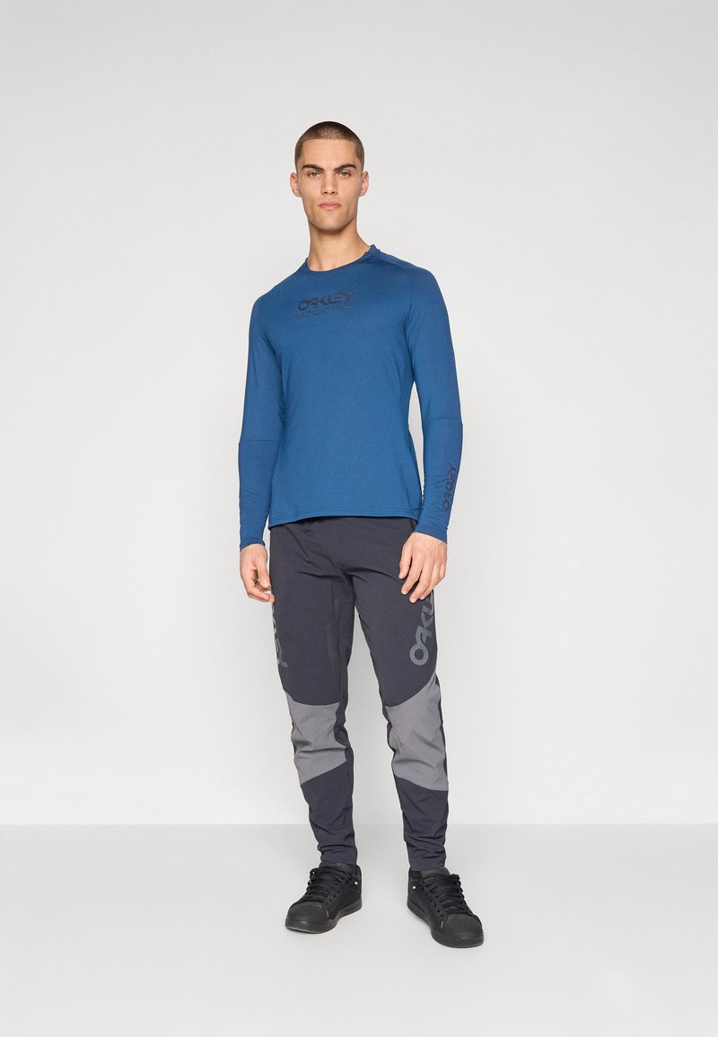 Camisa de manga larga azul con logo negro en el pecho, combinada con pantalones oscuros que presentan detalles y marca en gris. Tela texturizada, ajuste atlético.