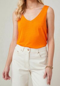 Top arancione senza maniche con scollatura a V, realizzato in tessuto morbido. Abbinato a pantaloni off-white a vita alta, evidenziando uno stile casual.