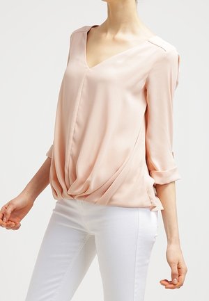 Vrouw draagt een lichtroze blouse met lange mouwen en een V-hals en witte aansluitende broek, poseert met licht gebogen armen.