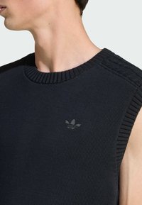 Crna bezrukavna pletena jumperica s rebrastim izrezom, koja ima suptilan tamni logo na prsima i teksturalne detalje na ramenima.