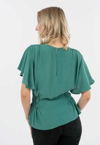 Blusa teal con maniche corte fluide, dettaglio in vita arricciato, scollatura a girocollo e chiusura con oblò sul retro. Abbinata a jeans neri.