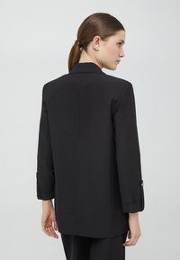 Czarna marynarka o dopasowanym kroju, z wyraźną konstrukcją, długimi rękawami i detalami na mankietach. Gładka tekstura o minimalistycznym stylu, bez widocznych wzorów.