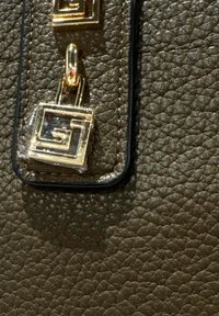 Primo piano di pelle marrone scuro texture con dettaglio di lucchetto geometrico color oro attaccato al bordo di una tracolla.