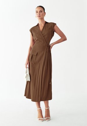 Woman wearing sleeveless brown wrap dress with pocket, holding small white handbag, and wearing white high-heeled sandals against white background.