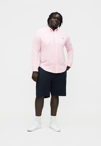 Mann som står iført en lyserosa skjorte med knapper, marineblå kortbukser til knærne, hvite sokker og hvite joggesko mot en ensfarget bakgrunn.