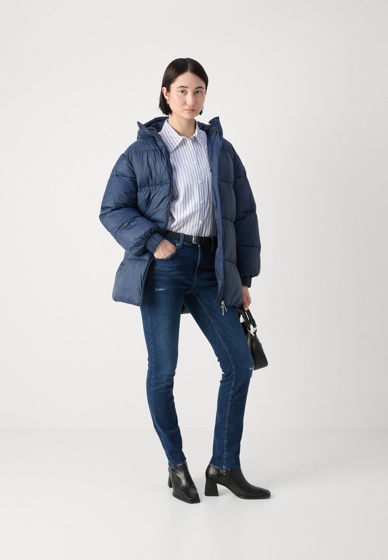 Chaqueta de plumas azul marino con capucha, camiseta blanca a rayas, vaqueros ajustados azul oscuro y botas negras de tobillo. Pequeño bolso negro con correa.