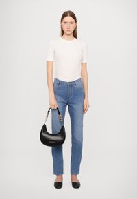 Črna usnjena hobo torba z zlatim verižnim dodatkom, ki jo drži oseba v beli oprijeti majici in modrih kavbojkah, ki stoji na enotni ozadju.