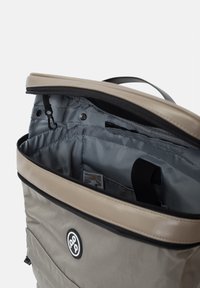 Harmaa kangaskassi, jossa on vaaleanruskea yläosa, vetoketjulla avautuva pääsylkä ja sisätaskut. Etuosassa on näkyvä logopatch. Pintamateriaali vaihtelee karkeista sileisiin.