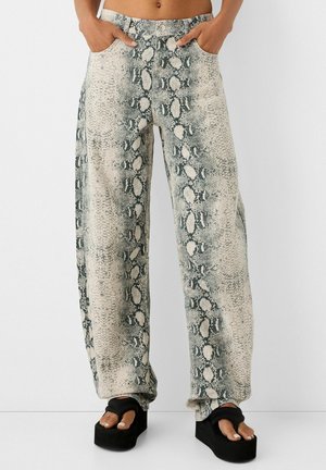 Vrouw draagt een broek met hoge taille en losse pasvorm, bedrukt met een grijs-witte slangenhuidprint, en zwarte platformsandalen met open tenen en enkelbandjes.
