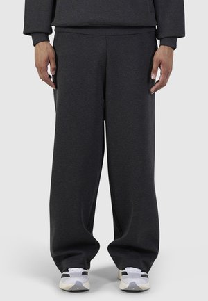 Onderlichaam van een persoon die losse donkergrijze broek en witte sneakers met zwarte en beige accenten draagt tegen een effen achtergrond.