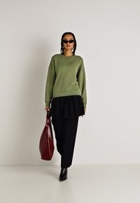 Suéter verde sobre una blusa negra de capas, combinado con pantalones negros y botines de punta. Sostiene un bolso rojo brillante. Texturas suaves.