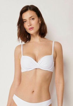 Junge Frau mit schulterlangem braunem Haar, die ein weißes Bikinioberteil und -unterteil vor einem schlichten, hellen Hintergrund trägt.