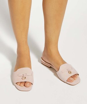 Pieds portant des sandales légères rose clair à bout ouvert, de type slip-on, avec de petits charms décoratifs dorés sur fond blanc.