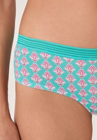 Slips bikini turchesi con un motivo floreale rosa indossati sui fianchi e sul torso inferiore di una persona, su uno sfondo semplice.