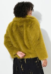 Faux fur jakke i sennepsgul, med en firkantet form, brede ærmer og krave. Båret over en sort nederdel med guld accenter.