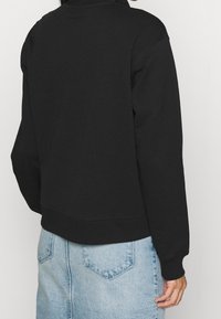 Svart sweatshirt med hög krage, ribbade muddar och en avslappnad passform. Sammanställt med en ljusblå denimkjol som visar en bakficka.