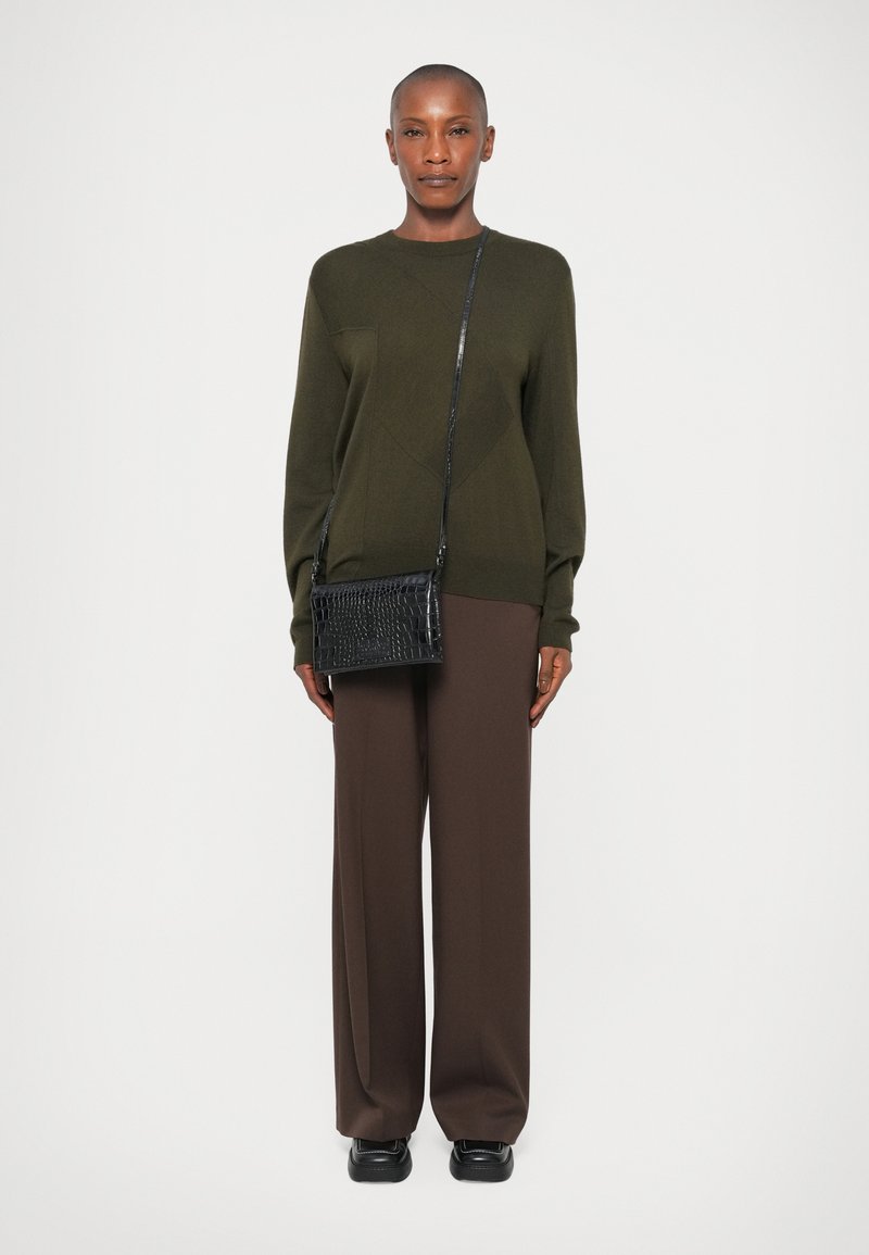 Maglione verde scuro con un motivo testurizzato, abbinato a pantaloni larghi marroni. Una borsa a tracolla nera con motivo coccodrillo completa l'outfit.