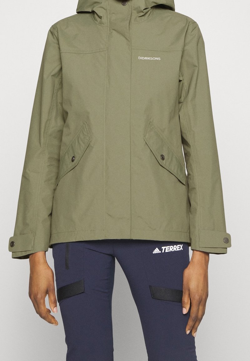 Veste à capuche verte avec poches avant boutonnées, portée avec un pantalon Adidas Terrex bleu marine.