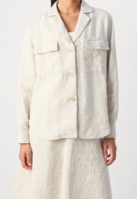 Camicia button-up in lino beige chiaro con colletto a scollo, due tasche frontali e maniche lunghe. Tessuto morbido con vestibilità rilassata.