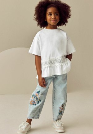 Hvid bomulds T-shirt med blonde kant nederst, parret med lyseblå jeans med blomsterbroderi. Hvide sneakers fuldender outfittet.