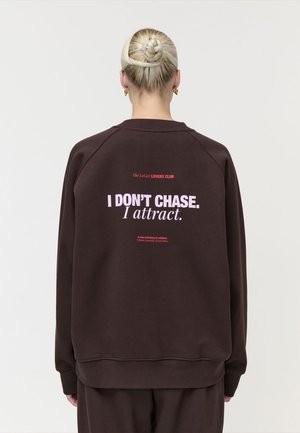 Persoon met blond haar in een gevlochten knot, die een donkerbruine trui draagt met daarop "I DON'T CHASE. I attract." in lichtroze letters op de achterkant.