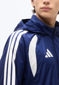 Άτομο που φοράει αθλητικό μπουφάν σκούρου μπλε με λευκές ρίγες στον ώμο και λευκό τρίγωνο λογότυπο στο στήθος, φερμουάρ κλειστό μέχρι τον λαιμό.