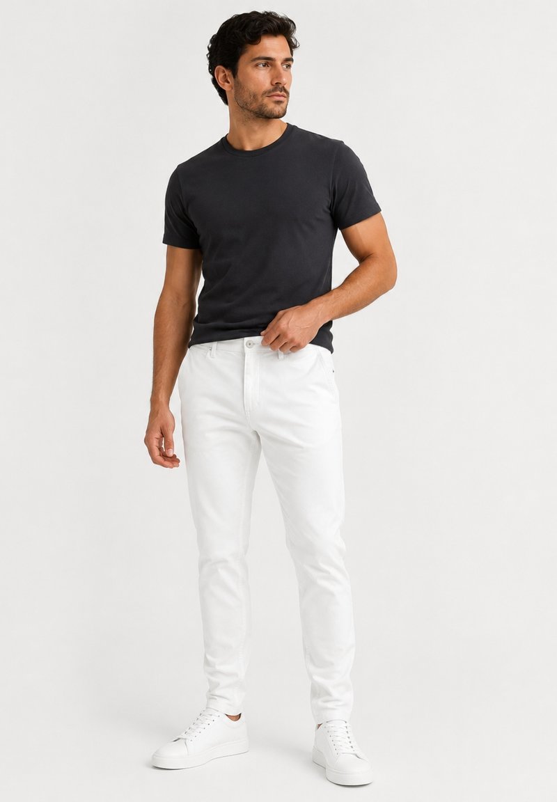 Man standing wearing a black short-sleeve t-shirt, white jeans, and white sneakers against a plain light background.