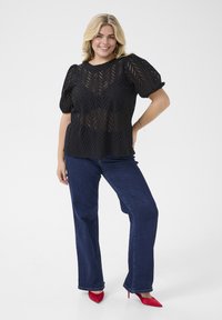 Black, short-sleeve top with a textured, cut-out geometric pattern. Paired with dark blue wide-leg jeans and red high-heeled shoes.