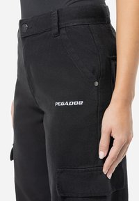 Black cargo pants made from sturdy fabric, feature a textured finish, pockets, a button closure, and "PEGADOR" embroidered logo on the side.