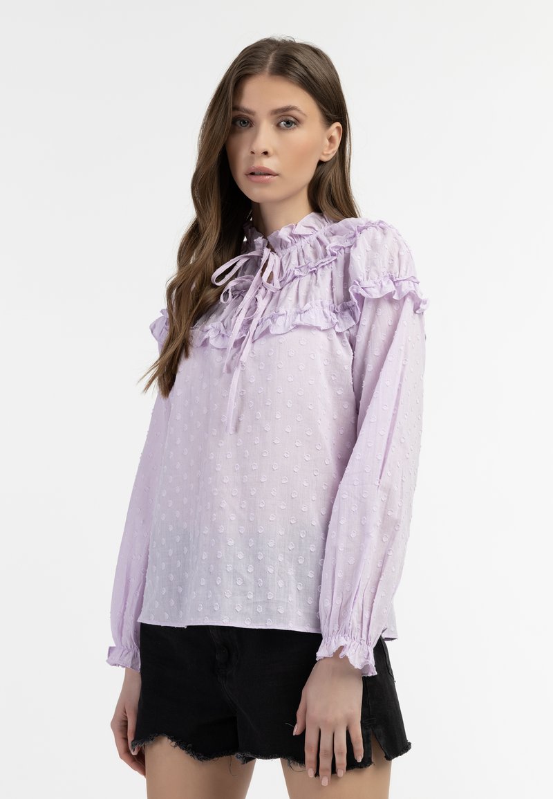 Lichtpaarse blouse met rimpelende halslijn en manchetten, voorzien van gemêleerde stippen en een gestrikte strikdetail. Draag met een zwarte shorts.