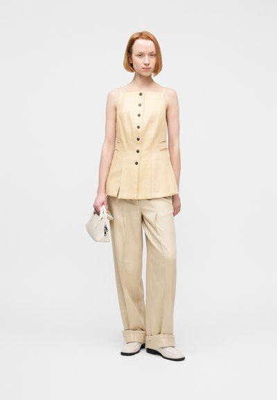 Γυναίκα με κοντά κόκκινα μαλλιά που φοράει μπεζ αμάνικο τοπ με κουμπιά, παντελόνι με φαρδιά πόδια, λευκά παπούτσια και κρατάει μικρή λευκή τσάντα.