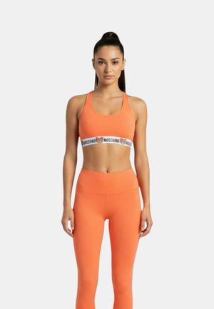 Donna con lunghi capelli scuri raccolti in una coda alta, indossa un reggiseno sport arancione e leggings abbinati, in piedi davanti a uno sfondo chiaro e uniforme.