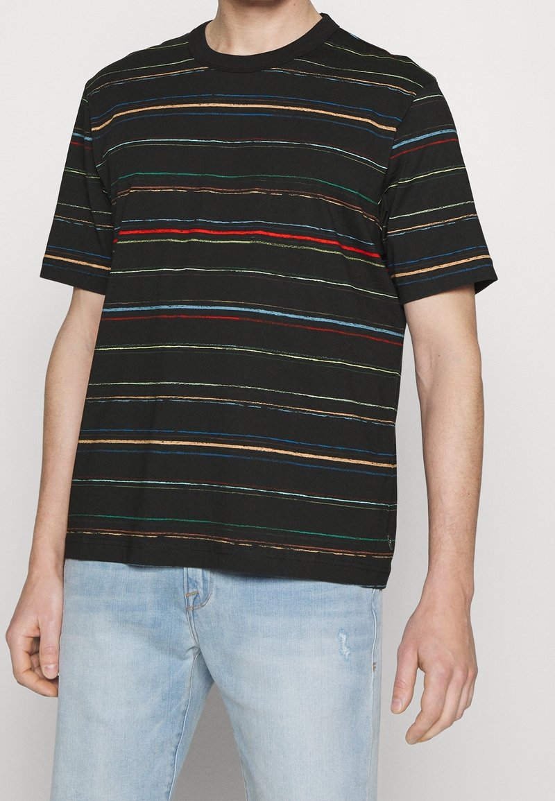 Man som bär en svart t-shirt med korta ärmar med tunna, multikolorade, horisontella ränder och ljusblå, slitna jeans.