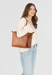 Braune Leder-Shopper-Tasche mit strukturierter Oberfläche, einem geräumigen Design und oberen Griffen, getragen von einer Person in einem cremefarbenen Pullover.