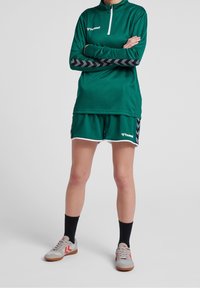 Top deportivo verde de manga larga con cremallera de un cuarto y detalles en forma de chevron negro, combinado con pantalones cortos a juego y zapatillas deportivas grises con detalles rojos.