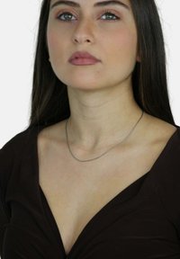 Srebrny łańcuszek na dekolcie, charakteryzujący się delikatnym, cienkim designem. Osoba nosząca ma długie ciemne włosy i głęboki brązowy top.