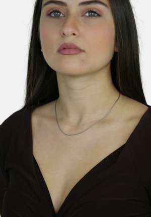 Collana in argento su un décolleté, caratterizzata da un design delicato e sottile. Chi la indossa ha lunghi capelli scuri e una maglia marrone scuro.