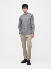 Jeune homme portant une chemise à rayures verticales à manches longues, un pantalon beige et des chaussures noires, debout sur un fond blanc uni.