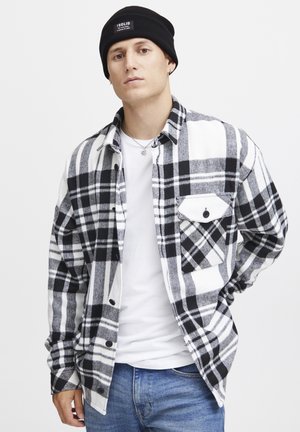 Camisa de franela a cuadros en blanco y negro con un bolsillo con botón, llevada sobre una camiseta blanca, combinada con un gorro negro. Ajuste casual, textura suave.