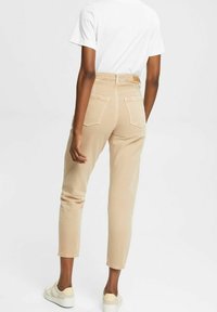 Jean taille haute beige avec une coupe fuselée, deux poches arrière et des ourlets retroussés. Porté avec un t-shirt blanc et des baskets de couleur claire.