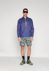 Tamsiai mėlynas ilgomis rankovėmis megztinis su geltonu užtrauktuku; mėlynai žali raštuoti šortai; juodos kojinės; daugių spalvų batai; dėvimi akiniai nuo saulės.